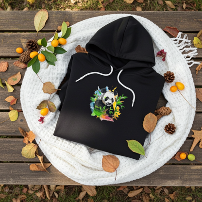 Black hoodie mockup featuring an embroidered panda with bamboo leaves and colorful splash accents on the chest, styled with autumn leaves, pinecones, and a cozy white knit blanket.