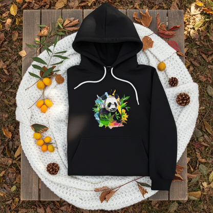 Black hoodie mockup featuring an embroidered panda with bamboo leaves and colorful splash accents on the chest, styled with autumn leaves, pinecones, and a cozy white knit blanket.