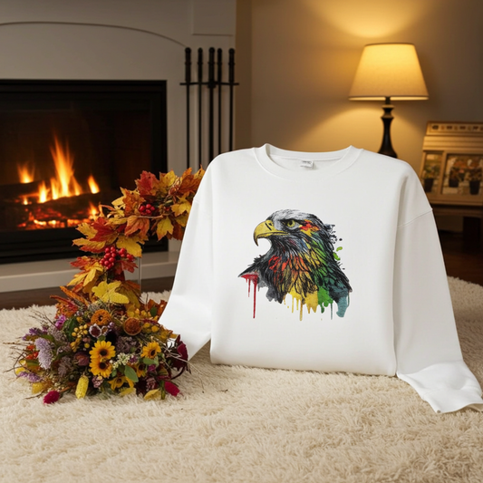 White sweatshirt displayed indoors near a fireplace, featuring a colorful splash-style eagle head embroidery on the chest, styled with autumn leaves and a bouquet on a soft carpet