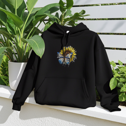 Black hoodie mockup featuring an embroidered blue butterfly and sunflower design on the chest, styled outdoors with lush green plants and a white fence backdrop.