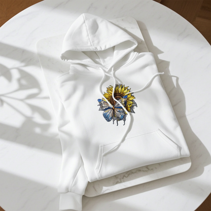 White hoodie mockup featuring an embroidered blue butterfly and sunflower design on the chest, laid flat on a round white table in bright natural light.