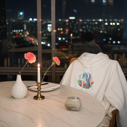 White hoodie mockup featuring an embroidered blue dragon with flowers on the chest, styled at a candlelit table with a city skyline view at night.