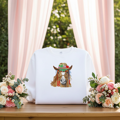 White sweatshirt displayed on wooden table with colorful boho-style horse embroidery wearing a decorated hat, surrounded by pastel floral arrangements and soft pink curtains in the background