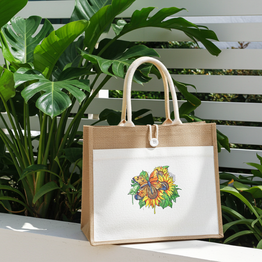 Natural jute tote handbag featuring a butterfly and sunflower embroidery design, displayed outdoors on a ledge with lush green tropical plants and a white slatted backdrop.