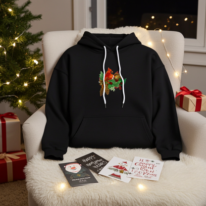 Black hoodie featuring a cardinal pair with pinecones embroidery design, styled on a cozy chair with string lights, holiday cards, a Christmas tree, and wrapped gifts nearby.