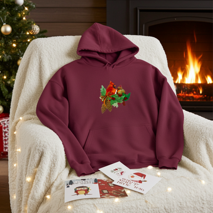 Burgundy hoodie featuring a cardinal pair with pinecones embroidery design, styled on a cozy armchair with string lights, holiday cards, a Christmas tree, and a lit fireplace in the background.
