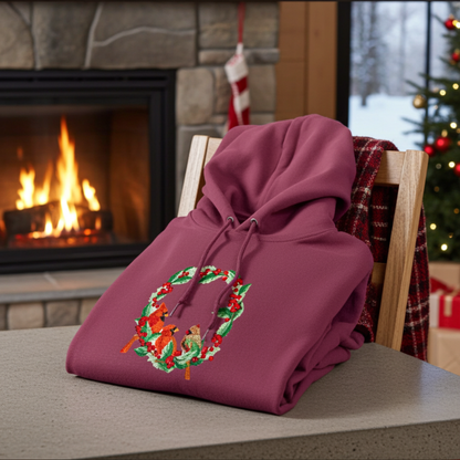 Burgundy hoodie featuring a cardinal wreath embroidery design, folded on a table with a plaid blanket, snowy window view, and a lit fireplace in the background.