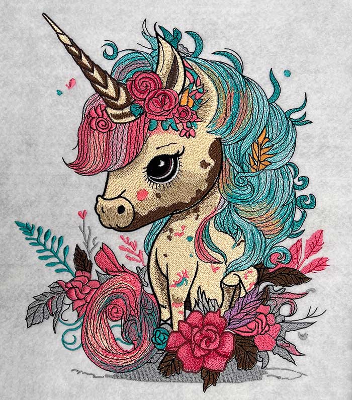 Close-up of an embroidered unicorn with a teal and pink mane, horn, and floral accents, stitched on a light fabric background.
