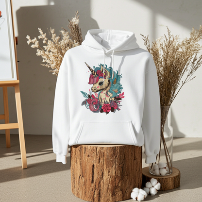 White hoodie mockup featuring an embroidered floral unicorn design on the chest, styled on a wooden stump with dried pampas grass and cotton stems against a wood wall.