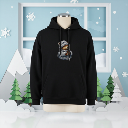 Black hoodie mockup featuring an embroidered bear in a knit hat and scarf holding a warm drink, displayed on a mannequin in a winter-themed scene with snowflakes, paper mountains, and a snowy window.