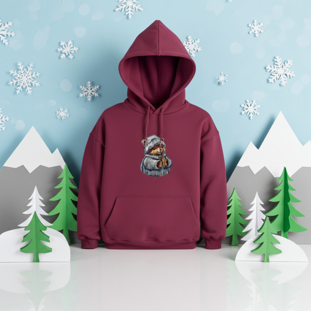 Burgundy hoodie mockup featuring an embroidered bear in a knit hat and scarf holding a warm drink, styled in a winter-themed scene with snowflakes and paper mountains.