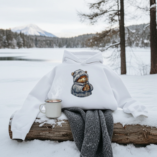 White hoodie mockup featuring an embroidered bear in a knit hat and scarf holding a warm drink, styled outdoors on a snowy log with a mug and scarf by a winter lake.
