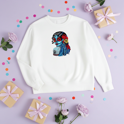 White sweatshirt laid flat on a lavender background with confetti and gift boxes, featuring a colorful sugar skull muse embroidery portrait with red and blue details on the chest