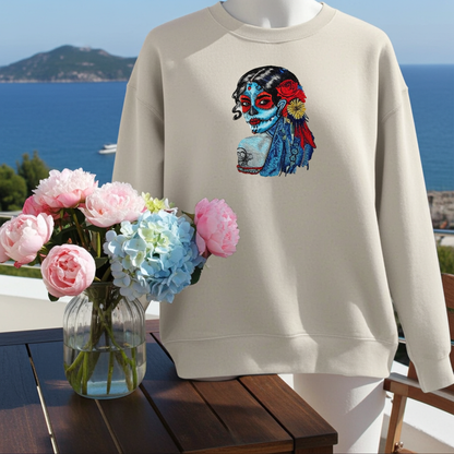 Mint green sweatshirt displayed on a balcony with ocean view, featuring a colorful sugar skull muse embroidery portrait with red and blue details, styled with a vase of pink and blue flowers on a wooden table