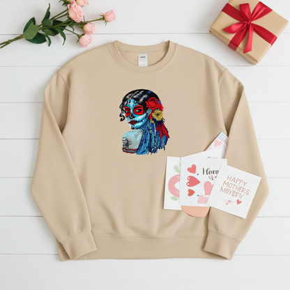 Soft beige sweatshirt laid flat on a white wooden background, featuring a colorful sugar skull muse embroidery portrait with red and blue details, styled with greeting cards, roses, and a wrapped gift