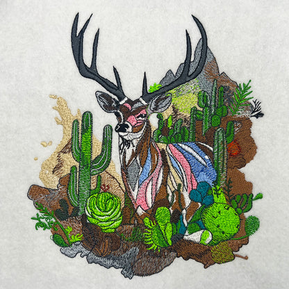 Close-up of an embroidered deer with large antlers standing among cacti and desert plants, stitched on a light fabric background.