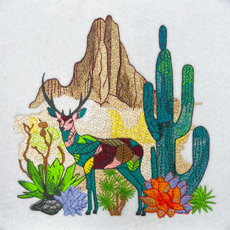 A jute tote bag with desert-themed embroidery including a deer, cacti, and flowers against a mountainous backdrop.