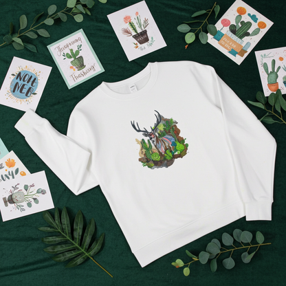 White sweatshirt laid flat on a dark green background with leafy branches and cactus-themed cards, featuring a desert prism deer embroidery design with antlers, cactus, and colorful landscape details on the chest