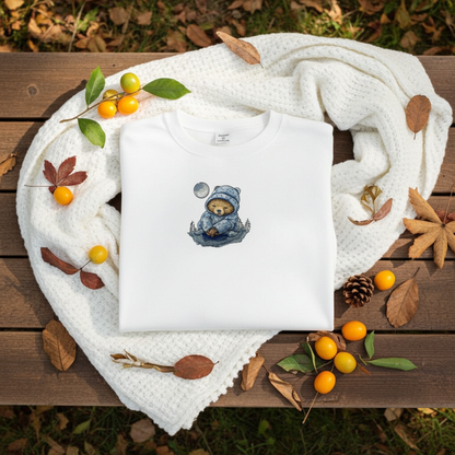 White sweatshirt folded on a chunky knit blanket outdoors, featuring a moonlit sloth embroidery in a cozy hat, styled with autumn leaves, pinecones, and small yellow berries on a wooden surface