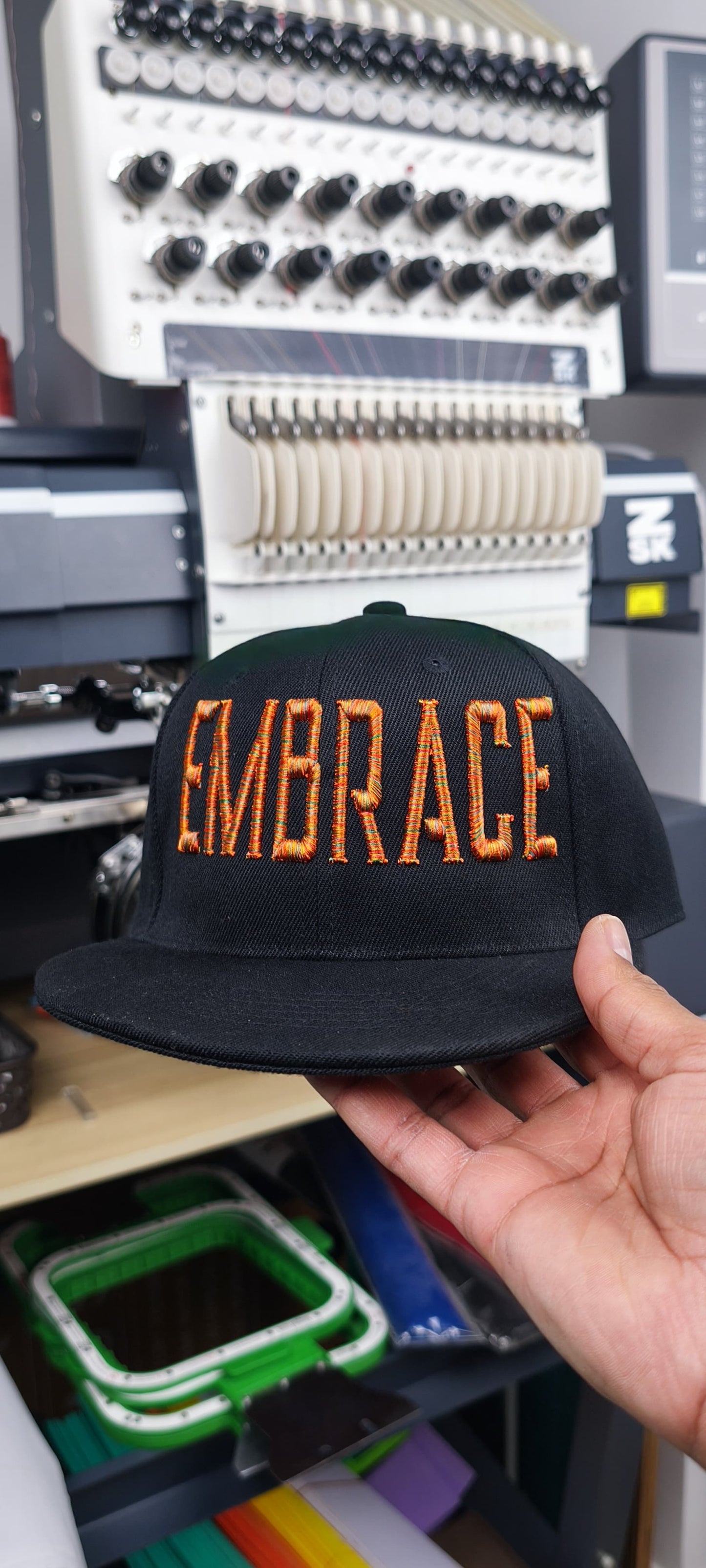 Black cap featuring “EMBRACE” in bold orange 3D puff embroidery lettering, held in hand in front of an embroidery machine in a studio setting, original EmbroMania work.