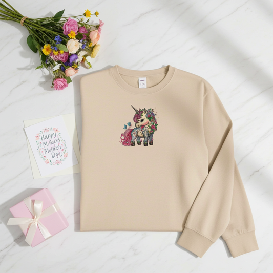 Soft beige sweatshirt laid flat on a white marble background, featuring an embroidered floral unicorn design on the chest, styled with a bouquet of flowers, a gift box, and a Mother’s Day card