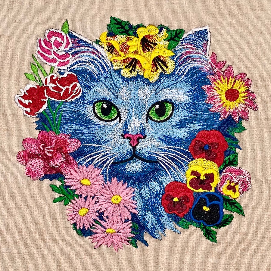 A tote bag with an embroidered design of a cat surrounded by colorful flowers on a beige background.
