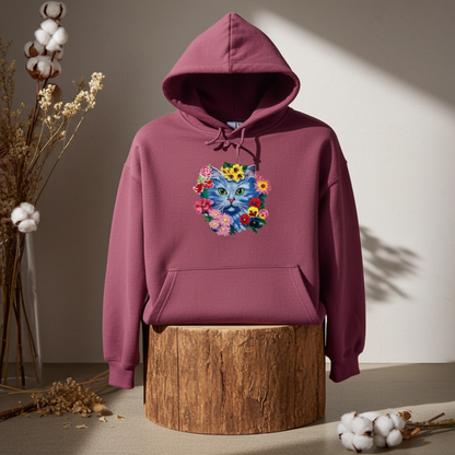 Burgundy hoodie mockup featuring an embroidered blue cat face surrounded by colorful flowers on the chest, displayed on a wooden stump with dried florals and cotton stems in warm light.