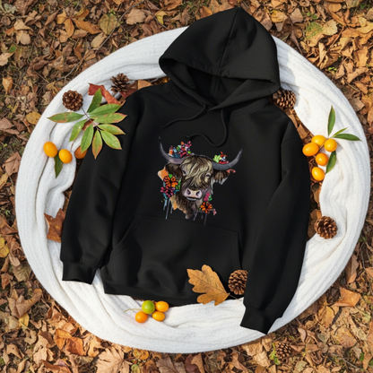 Black hoodie mockup featuring an embroidered highland cow with long horns and colorful wildflowers on the chest, styled outdoors on a white blanket over autumn leaves with pinecones, green foliage, and small orange berries.