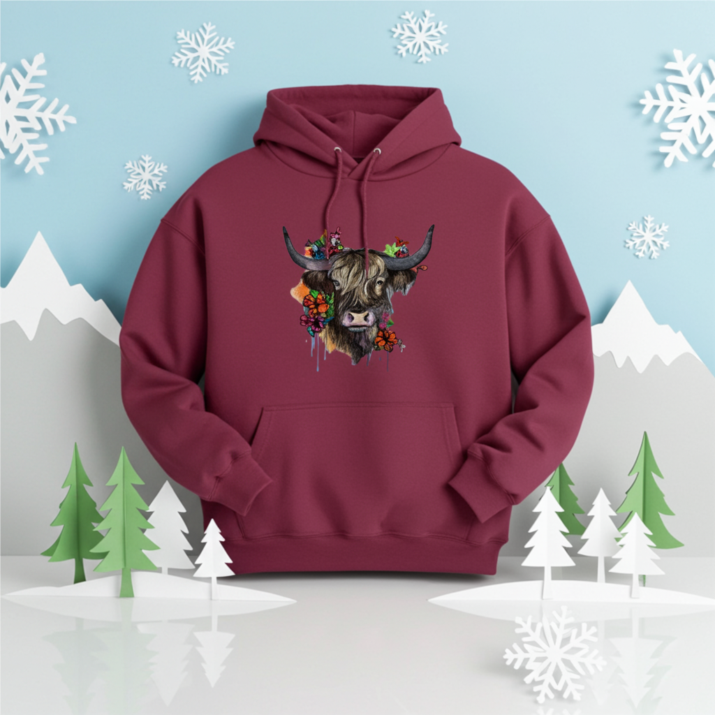 Burgundy hoodie mockup featuring an embroidered highland cow with long horns and colorful wildflowers on the chest, displayed in a winter-themed scene with snowflakes, paper mountains, and stylized pine trees.