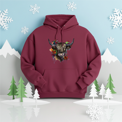 Burgundy hoodie mockup featuring an embroidered highland cow with long horns and colorful wildflowers on the chest, displayed in a winter-themed scene with snowflakes, paper mountains, and stylized pine trees.