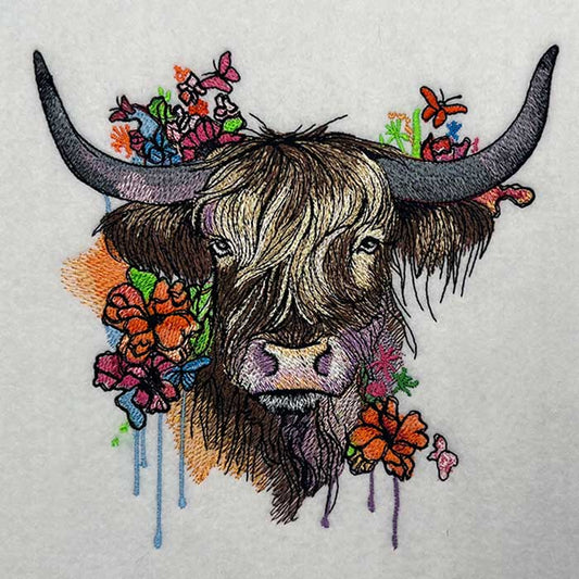 Highland cow embroidery design featuring a shaggy cow face with long horns, surrounded by colorful wildflowers and butterflies with paint-drip style accents on light fabric.