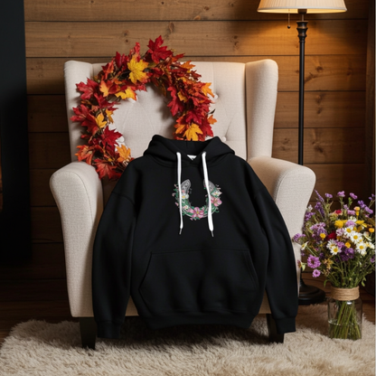 Black hoodie mockup with a floral embroidered horseshoe design on the chest, styled on an armchair with an autumn leaf wreath, warm lamp lighting, and a bouquet of flowers.
