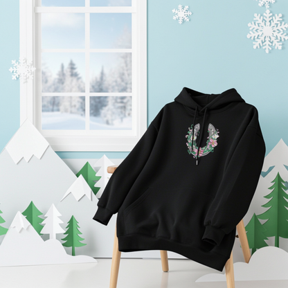 Black hoodie mockup featuring a floral embroidered horseshoe design on the chest, styled in a winter-themed room with snowflakes, paper mountains, and a snowy window backdrop.