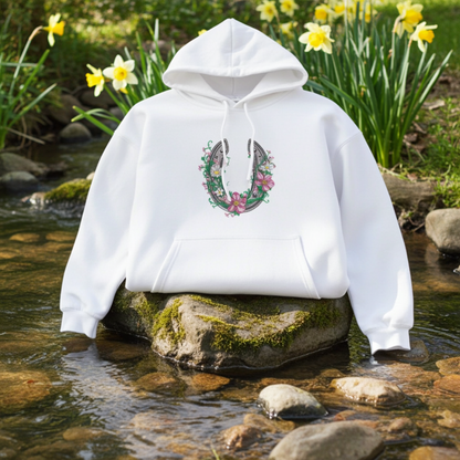 White hoodie mockup with a floral embroidered horseshoe design on the chest, styled outdoors on a mossy rock in a shallow stream with yellow flowers in the background.