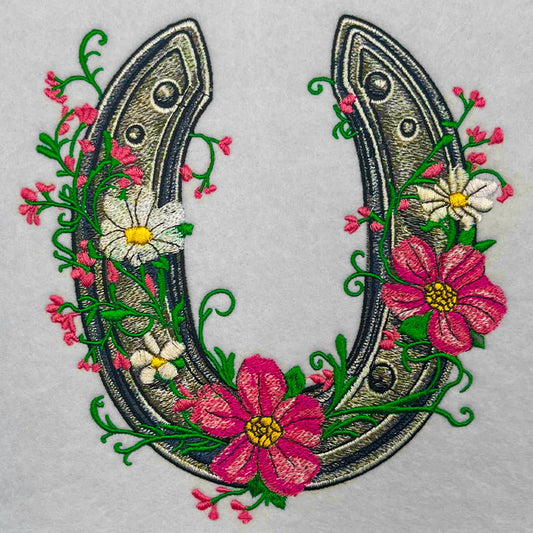 Close-up of an embroidered horseshoe decorated with pink and white flowers, green vines, and small pink buds on a light fabric background.