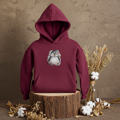 Burgundy hoodie mockup featuring an embroidered cute penguin with floral accents on the chest, displayed on a wooden stump against a textured wall with dried flowers and cotton stems.