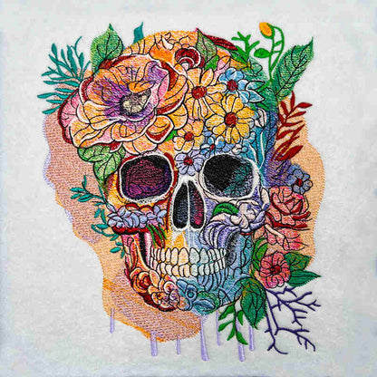 Floral skull embroidery design featuring a colorful skull covered with vibrant flowers and green leaves, detailed stitching with paint-drip accents on light fabric.