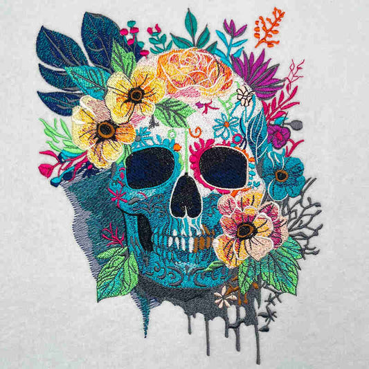 Floral skull embroidery design featuring a turquoise skull with colorful flowers and leaves, detailed stitching and paint-drip accents on light fabric.