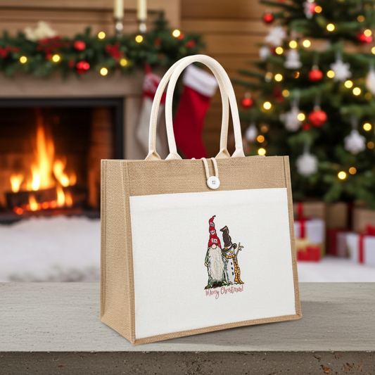 Natural jute tote bag featuring a gnome and snowman “Merry Christmas!” embroidery design, styled indoors with a decorated Christmas tree and a lit fireplace in the background