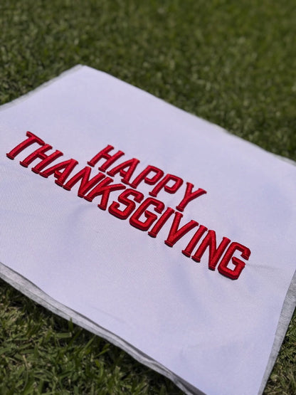 Red satin-stitch embroidery reading “HAPPY THANKSGIVING” in Metro Block lettering on white fabric, photographed on grass.