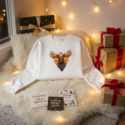 White sweatshirt folded on a fluffy rug with warm fairy lights, featuring a detailed moose embroidery with autumn leaves and orange accents, surrounded by wrapped gifts and holiday cards