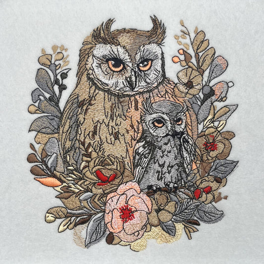 Detailed embroidery artwork of a mama owl and baby owl surrounded by woodland leaves and flowers in neutral tones with soft pink and red accents, original EmbroMania design.