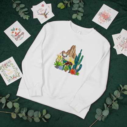 White sweatshirt laid flat on a dark green background with eucalyptus leaves and illustrated cards, featuring a geometric desert deer embroidery with cactus and rock details on the chest