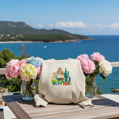 Mint green sweatshirt displayed on an outdoor table with ocean and mountain view, featuring a geometric desert deer embroidery with cactus and rock details, styled with vases of pink and white flowers in sunlight
