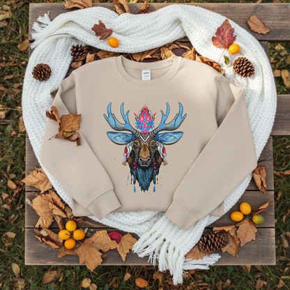 Soft beige sweatshirt folded on knitted white blanket outdoors, featuring vibrant blue antler moose embroidery with pink accents, surrounded by autumn leaves, pinecones, and yellow berries on wooden bench