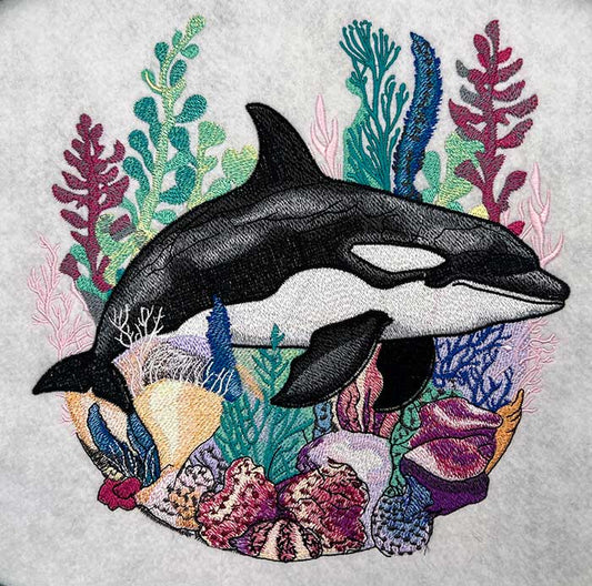 Embroidered orca whale swimming through a colorful coral reef with sea plants and shells in teal, pink, purple, and blue tones, original EmbroMania design.