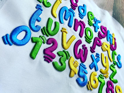 Colorful 3D puff embroidery letters and numbers in a playful kids-style font (A–Z and 0–9) stitched on white fabric in blue, green, purple, and yellow thread.