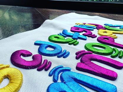 Angled close-up of colorful 3D puff embroidered letters and numbers in a playful kids-style font on white fabric, stitched in purple, blue, green, and yellow thread.