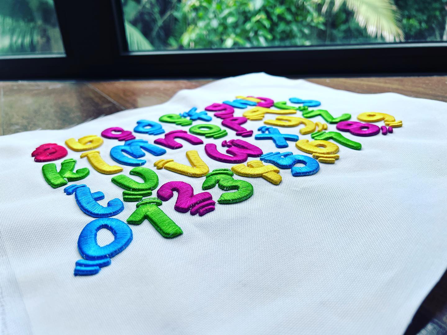 Colorful 3D puff embroidered alphabet letters and numbers arranged in a curved layout on white fabric, photographed on a table near a window with greenery outside.