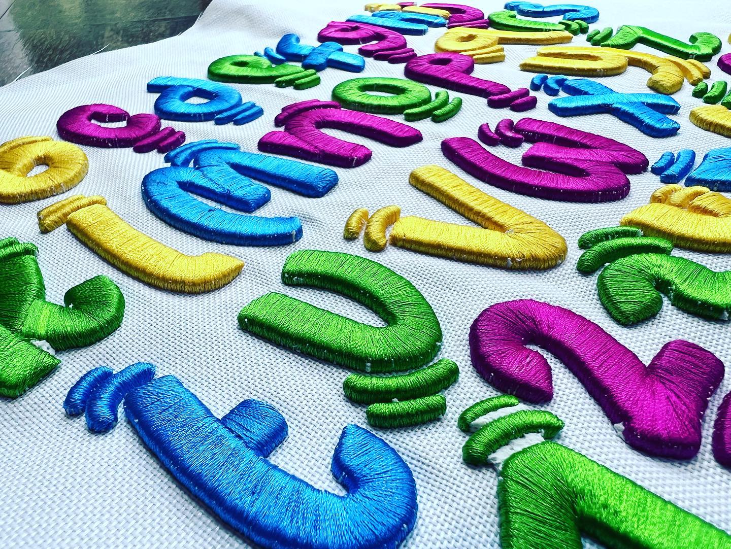 Wide close-up of colorful 3D puff embroidered alphabet letters and numbers spread across white fabric, stitched in bright blue, green, purple, and yellow thread.